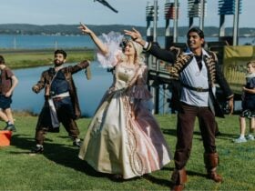 Three costumed performers in theatrical outfits strike dynamic poses near the water during an outdoo