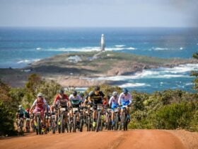 Mountain bike group racing up steep red gravel road, with coast and lighthouse in background