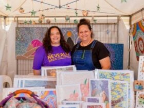 Two female Aboriginal artists stand behind their stall displaying authentic Aboriginal artworks.