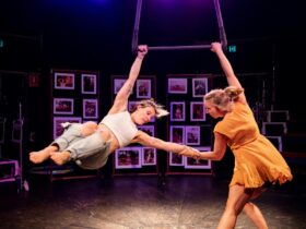 Two women hanging on a trapeze and holding hands in front of a wall of photos.