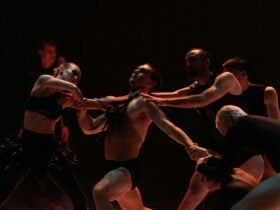 Dancers in dark costumes intertwine in a dramatic, low-lit stage scene, with one performer supported