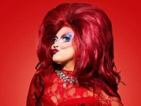 Woman in drag with large make up and red hair staring at the camera with a cheeky smile.